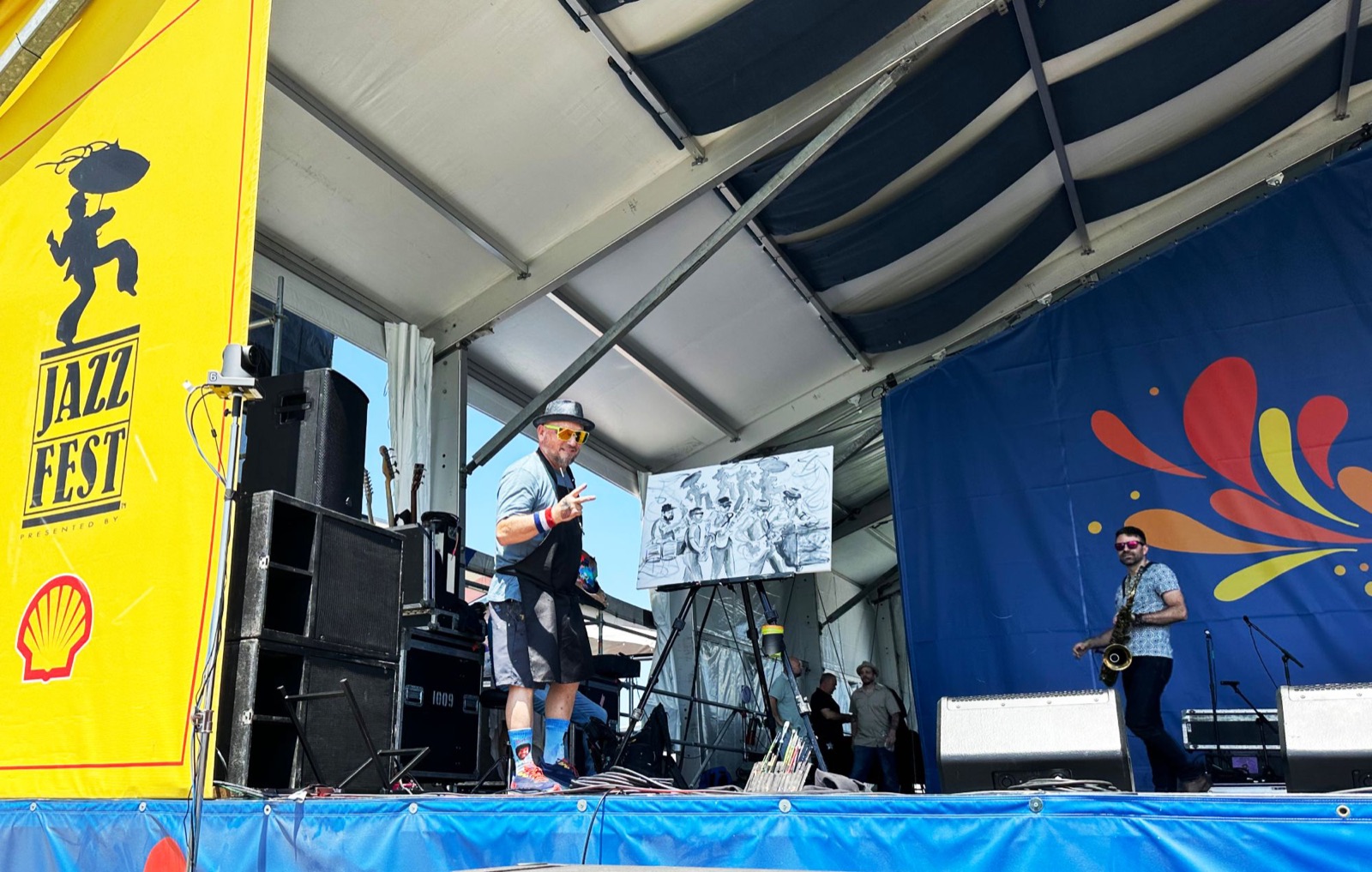 Frenchy on a Jazz Fest stage beside his easel, the band in black and white on the canvas, as a saxophonist plays