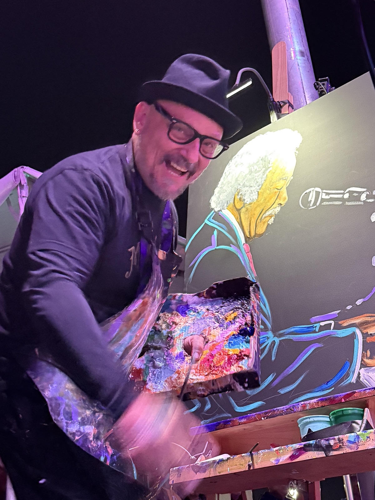 Frenchy grinning at his easel on the NOLA Funk Fest main stage with his Allen Toussaint painting behind him